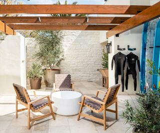 California casual style outdoor patio with oversized garden chairs surrounding a coffee table. Surfboards lean up against the wall, while wetsuits hang from a wooden beam of the pergola