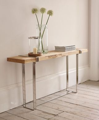 Plato Console Table with wooden top and wrapped in a steel frame, set against a pale wall with a vase of flowers, candle and books on top