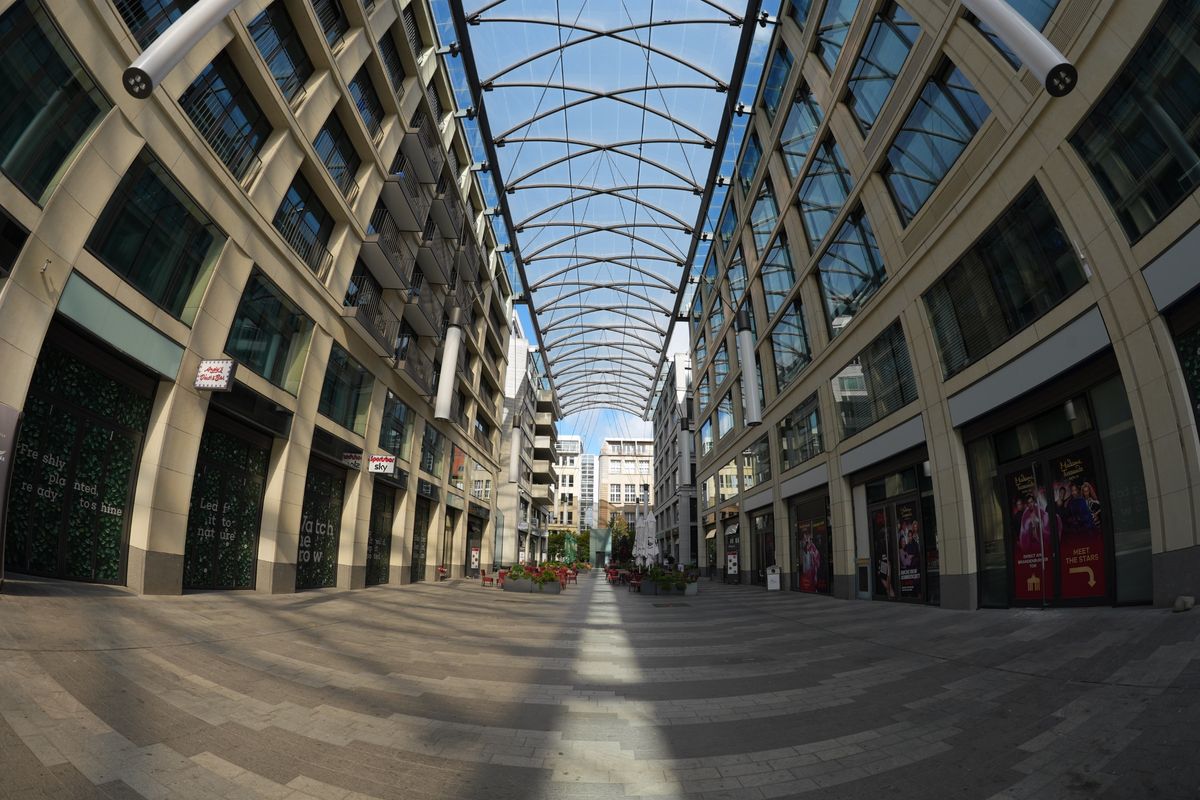 Ultra-wide view along a modern pedestrian courtyard in Berlin, with tall buildings and an arched glass canopy overhead.