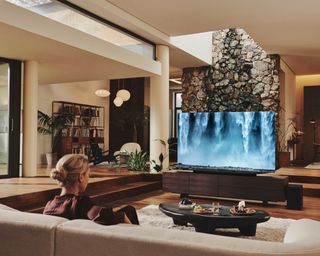 Samsung QN900B TV with soundbar and subwoofer sitting on dark wood TV stand opposite woman
