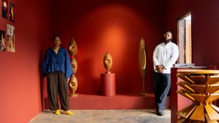 Two young designer, a woman, and a man, stand in front of their sculptural, drop-shaped wooden lamps in a red-painted gallery room.