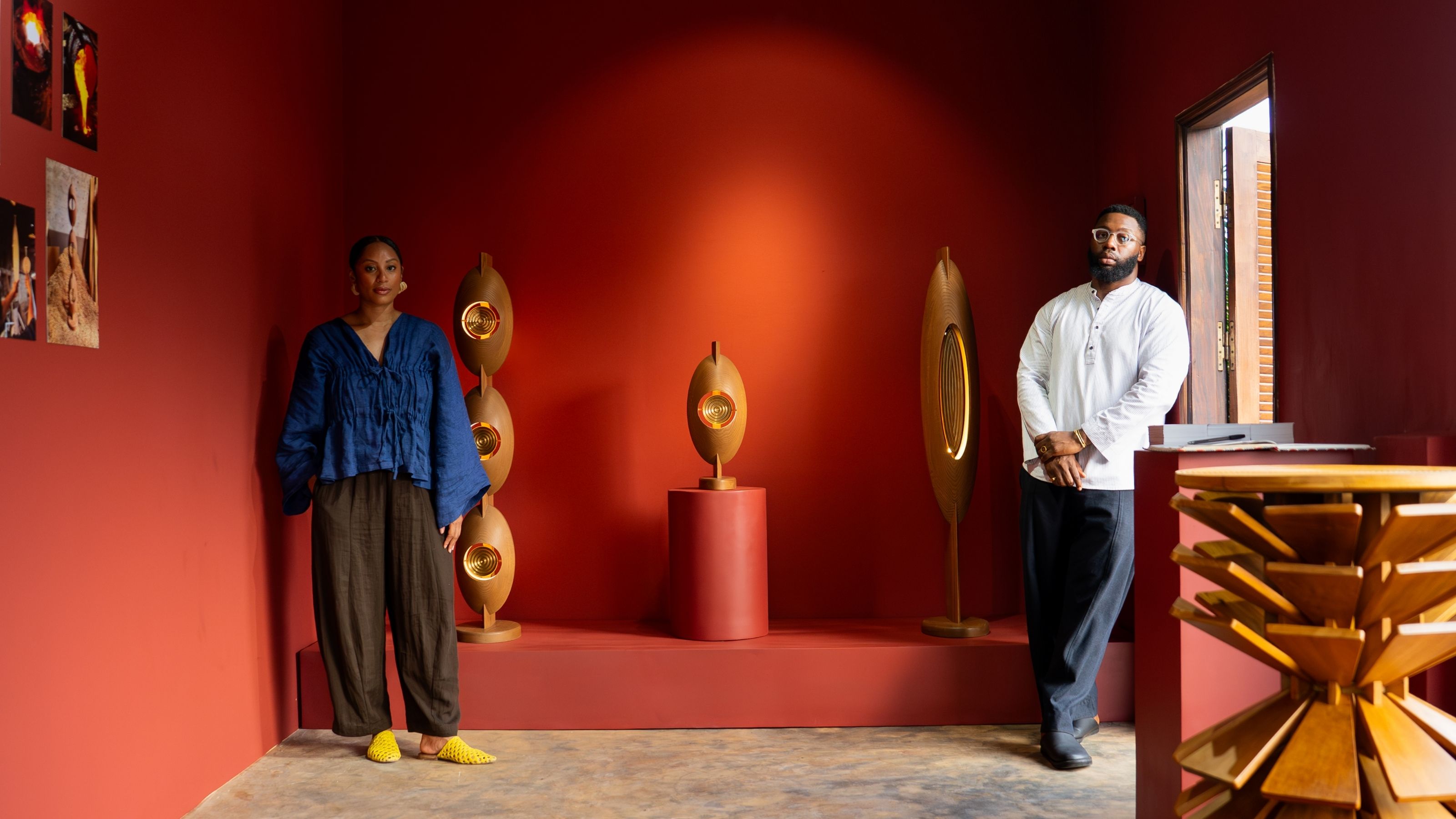 Two young designer, a woman, and a man, stand in front of their sculptural, drop-shaped wooden lamps in a red-painted gallery room.