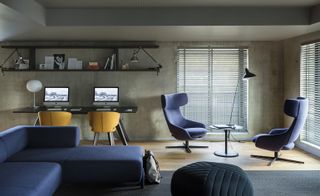 Workspace with a dark corner-sofa, two wing-back chairs and a coffee table, and a desk with two laptops in the background