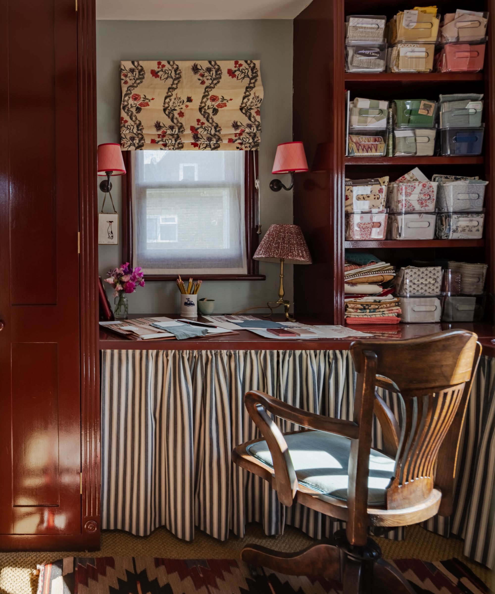 Small home office with red painted built-in cupboards, built-in desk with skirted fabric, and an open cupboard with plastic containers holding stationary
