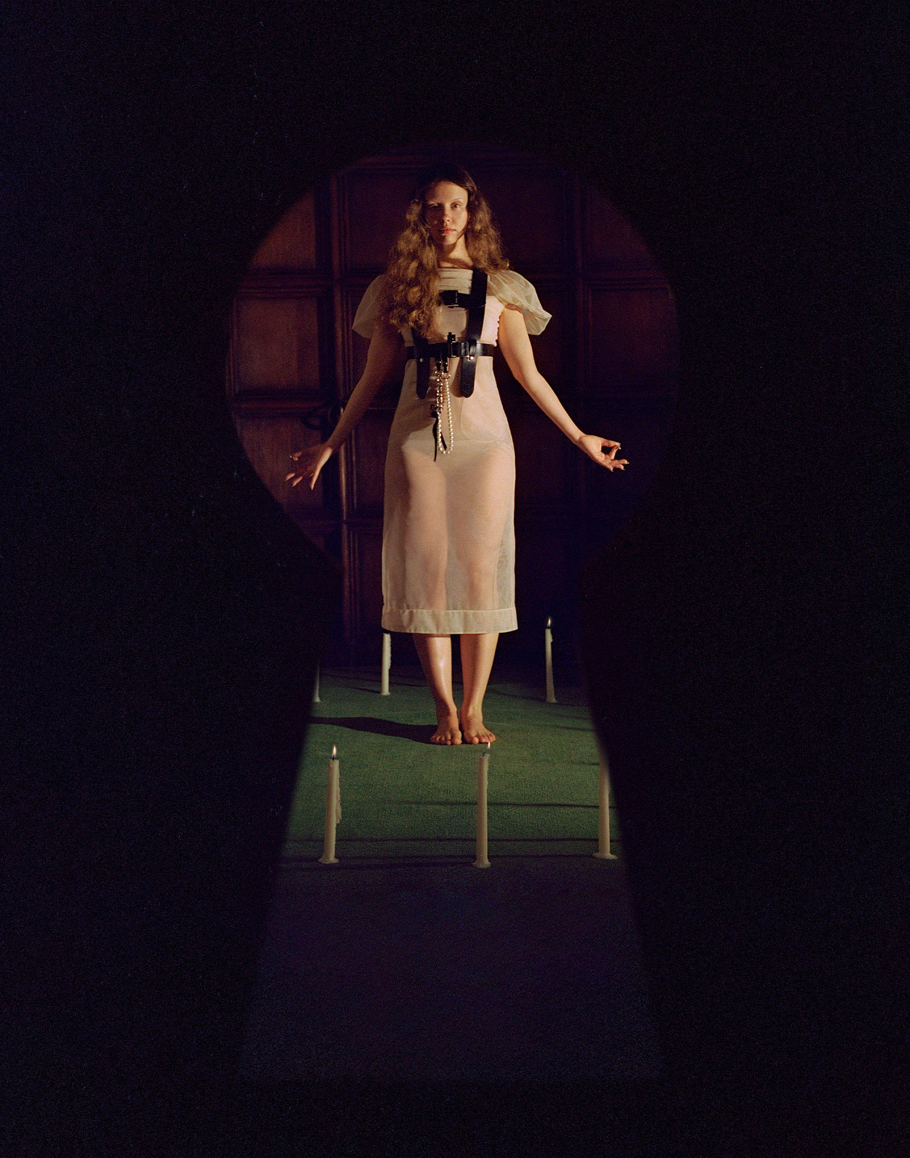 Mia Goth photo shoot for Who What Wear&#039;s October Cover Story. In this image, which as the perspective of looking through a keyhole, Mia is seen standing inside a circle of candles wearing a thin, sheer white dress with leather harness.