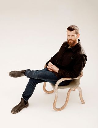 View of Matthew Day Jackson dressed in a brown jacket, blue jeans and brown boots sitting in his 'Kolho' chair prototype against a light coloured background