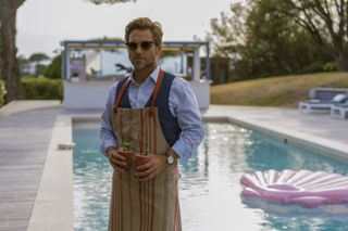 Harry (Jamie Bamber) stands in front of the swimming pool in the back garden of a French villa, wearing sunglasses and with an apron covering his smart shirt, waistcoat and trousers, holding two glasses of smoothies