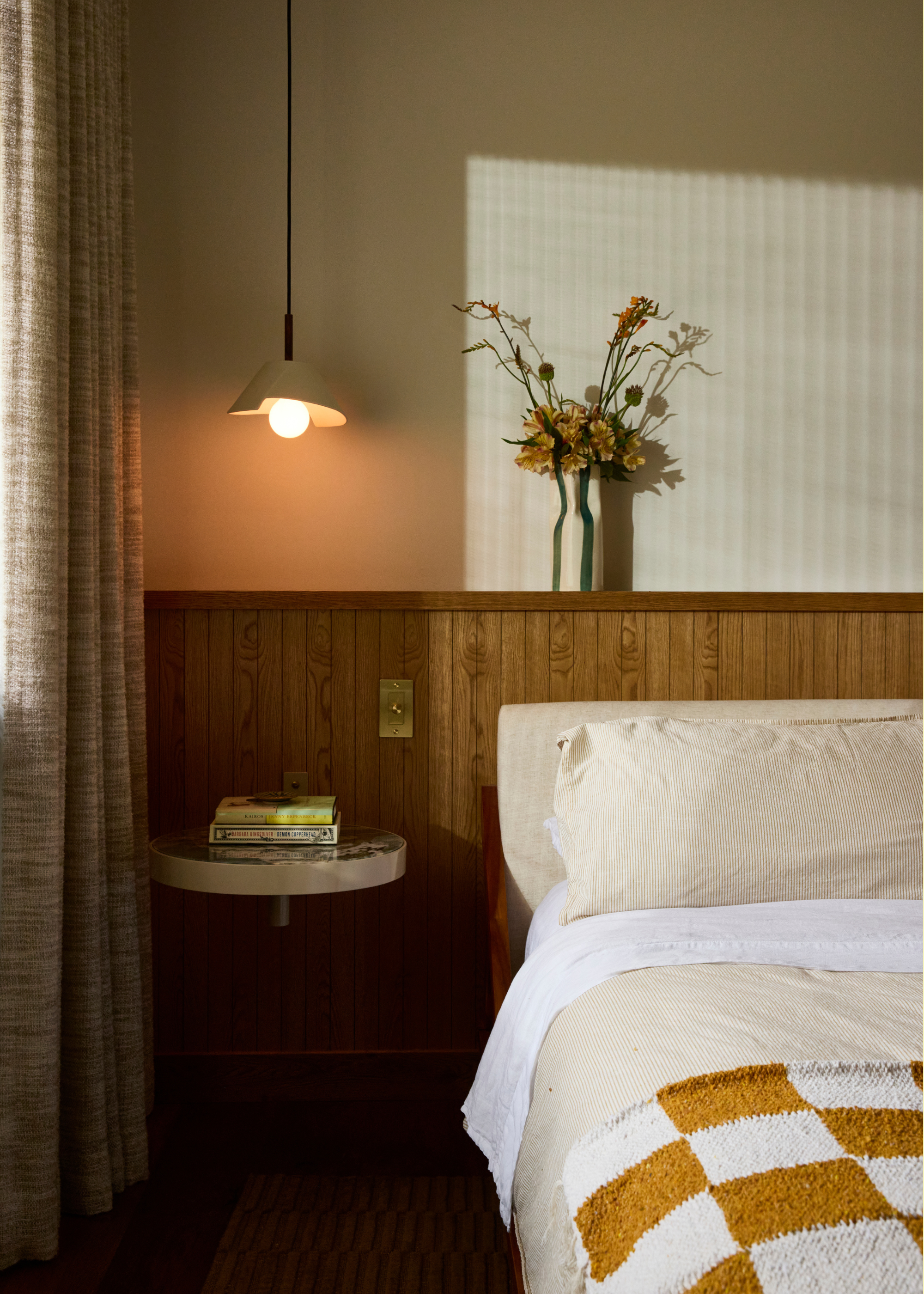 A white bedroom with a wood panelled headboard, a vase of flowers, a pendant lamp above a floating nightstand with a stack of books
