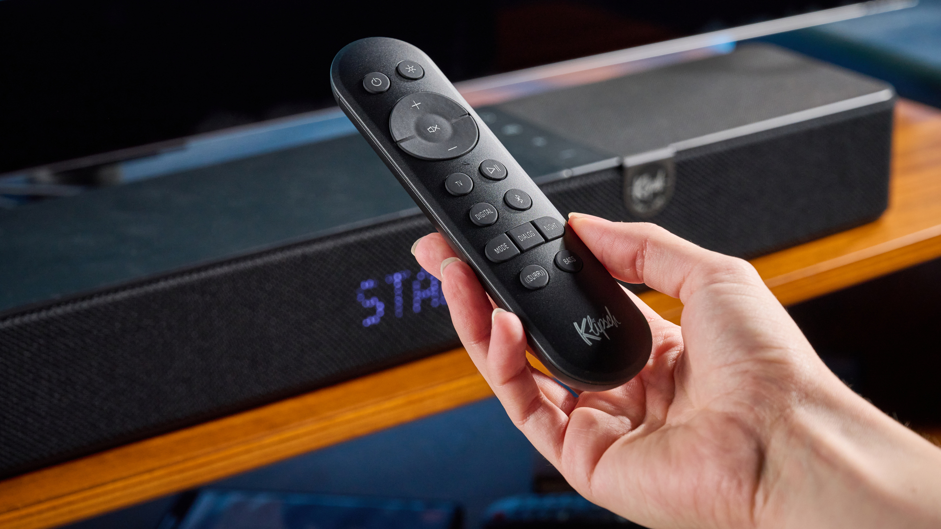 the klipsch flexus core 100 soundbar photographed on a wooden tv stand with a tv behind, showing the remote, buttons, and inputs