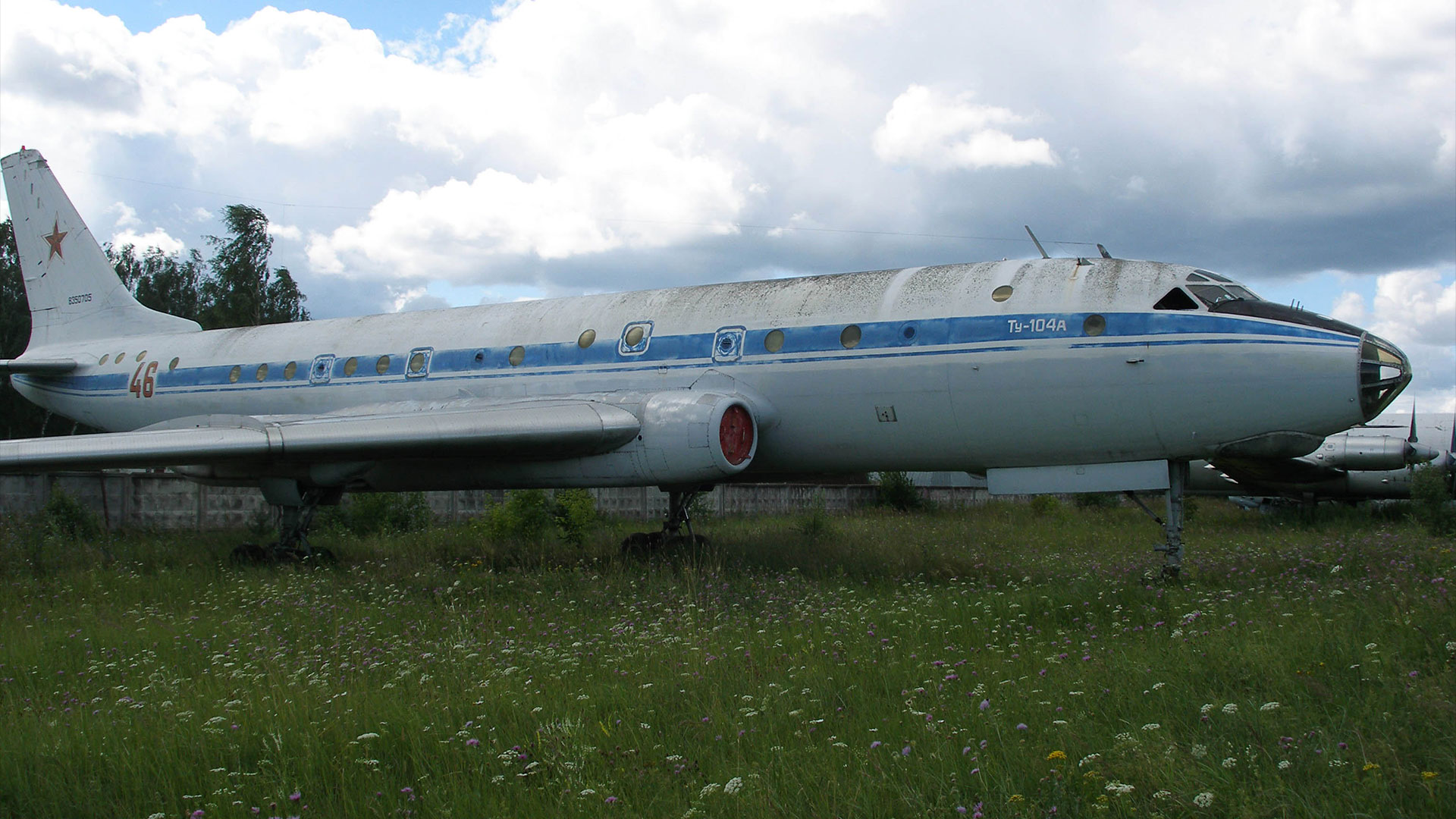 a jet aircraft with a white and blue livery sits in a grass field under a mostly cloud covered blue sky
