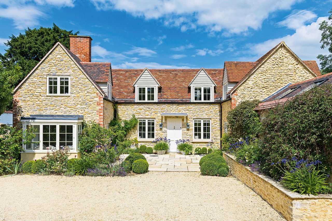 pale stone cottage with new wing white front door and flower beds in bloom and blue sky