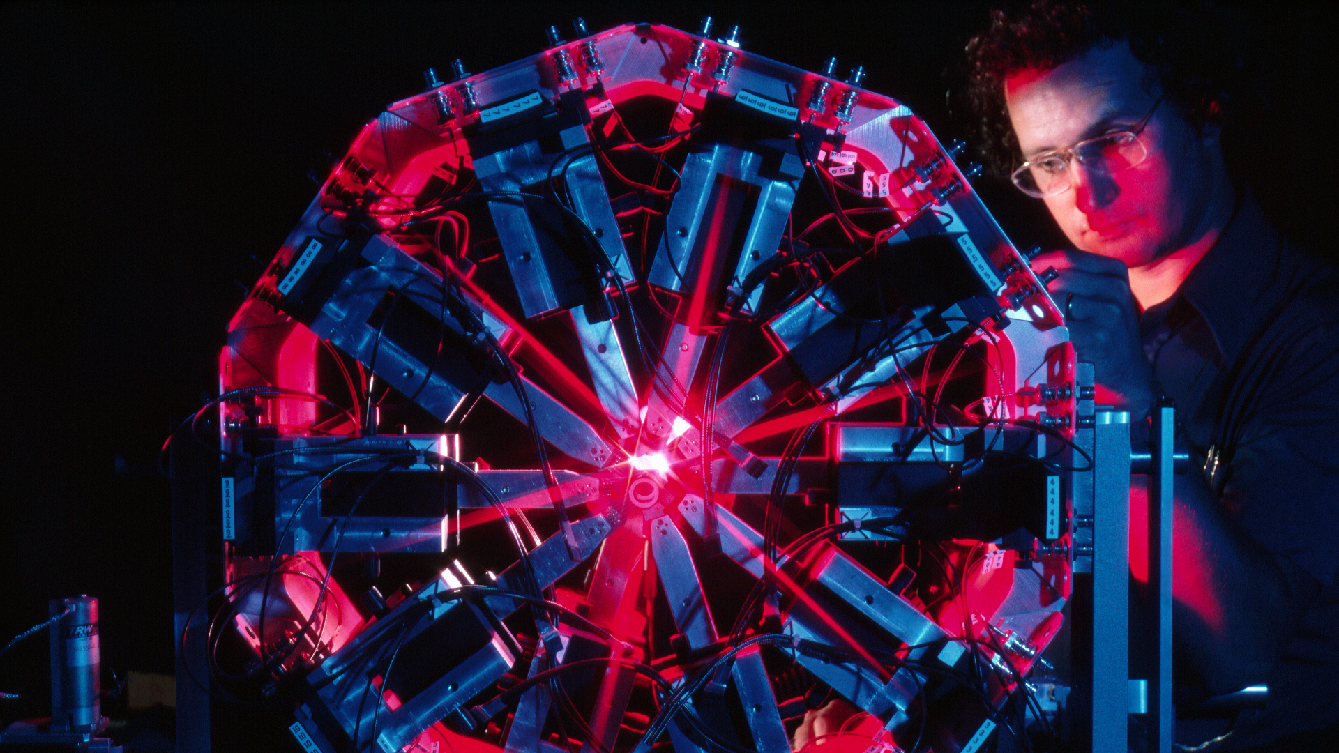 A man with glasses and a blue shirt stands behind a Ferris-wheel looking contraption with red and blue lighting in a dark room