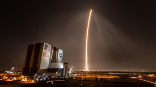 a streak of light stretches from the distant horizon up into the sky beside a large white building with an american flag and a round blue circle with the word "NASA" on it