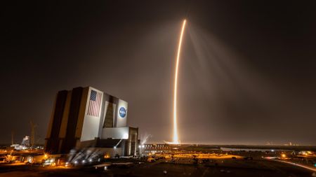 a streak of light stretches from the distant horizon up into the sky beside a large white building with an american flag and a round blue circle with the word "NASA" on it