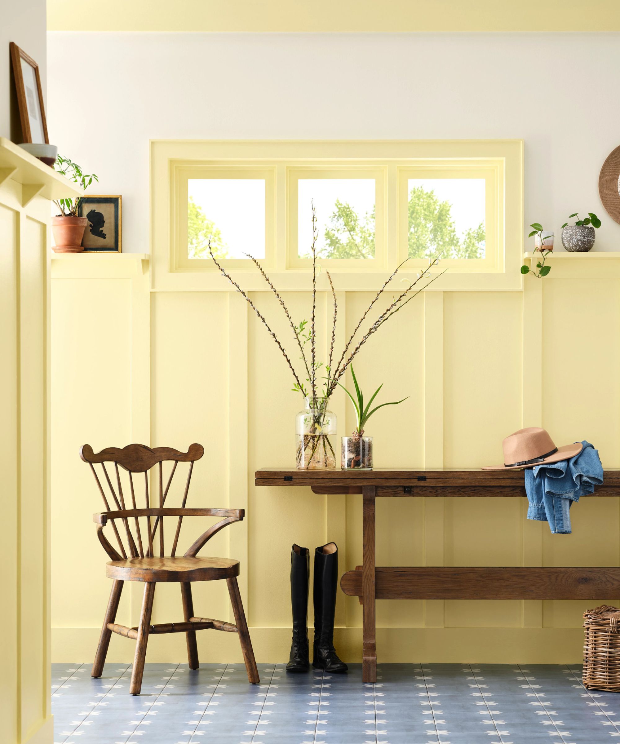 a panelled entryway painted in a sunshine yellow with a rustic wooden bench, vintage wooden chair, boots, and vases of foliage
