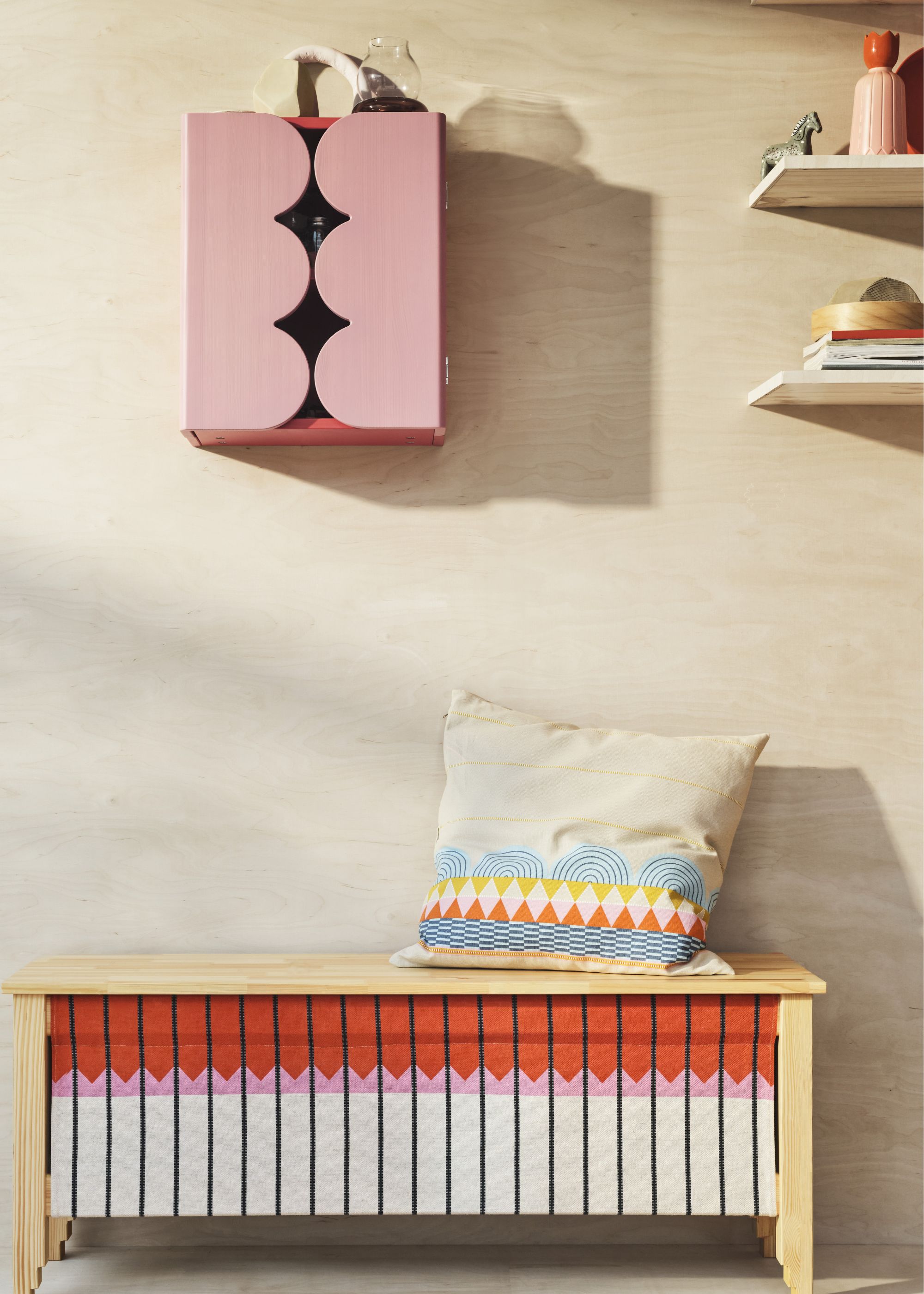 A pink scallop colored cabinet with a bench beneath it as well as a pillow on top. There are also floating shelves on the right side of the room with accessories on top