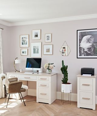 A bedroom with lilac walls with a white and gold gallery wall, a beige desk with a lamp, a computer, and flowers, a brown leather chair, and a green cacti plant next to it in a white pot
