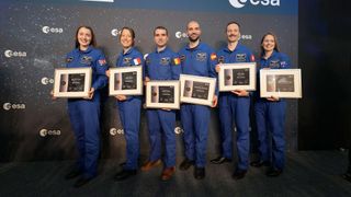 six people in blue flight suits stand on a stage, each one holding a plaque