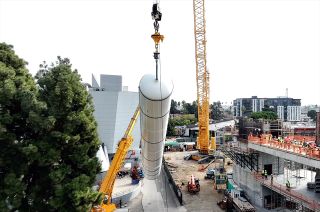 two cranes hoist a large white cylindrical rocket motor into place in an unfinished building.