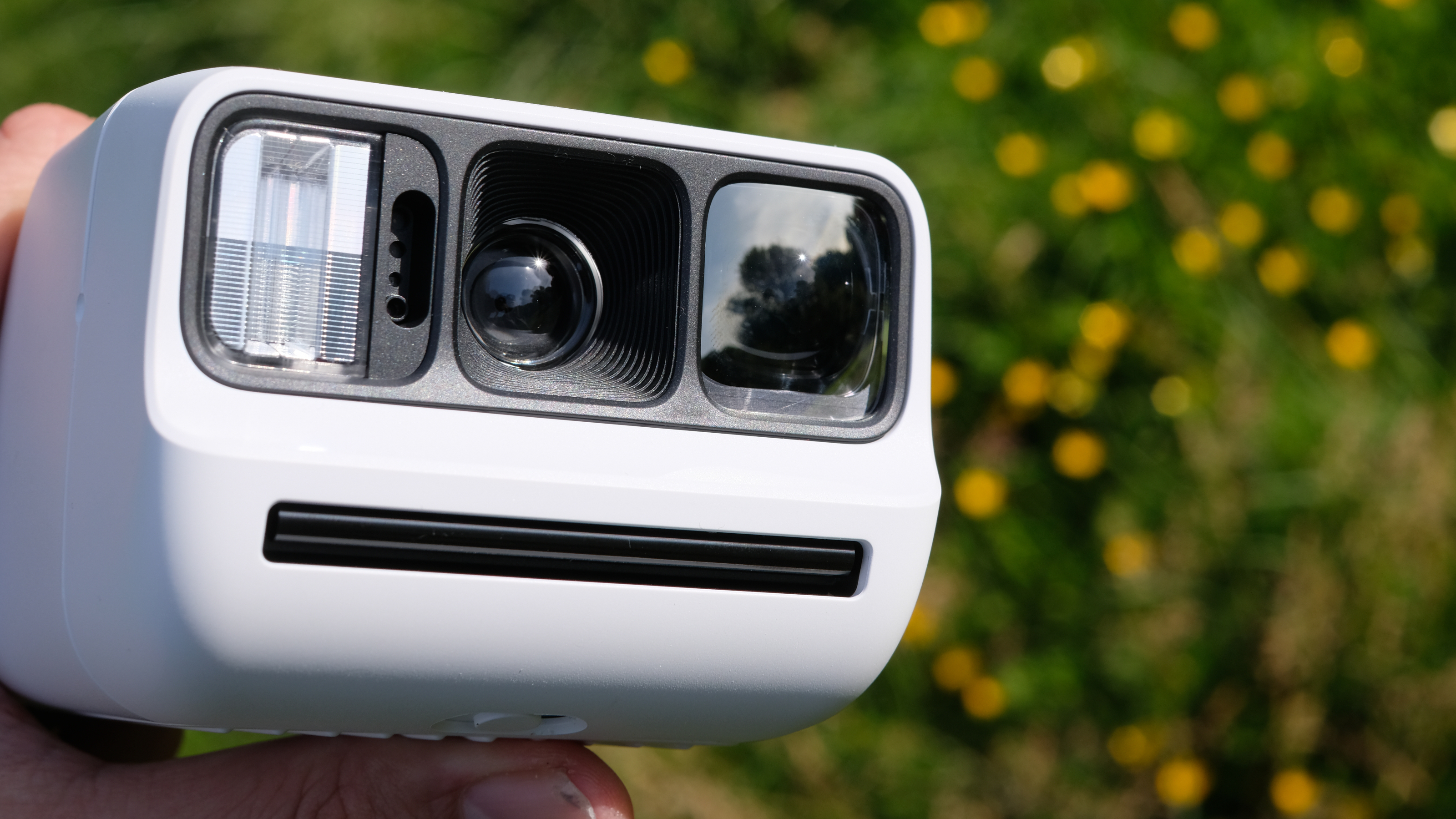 Photograph of man's hand holding Polaroid Go Gen 3 camera in white, with close front view of flash, lens and viewfinder