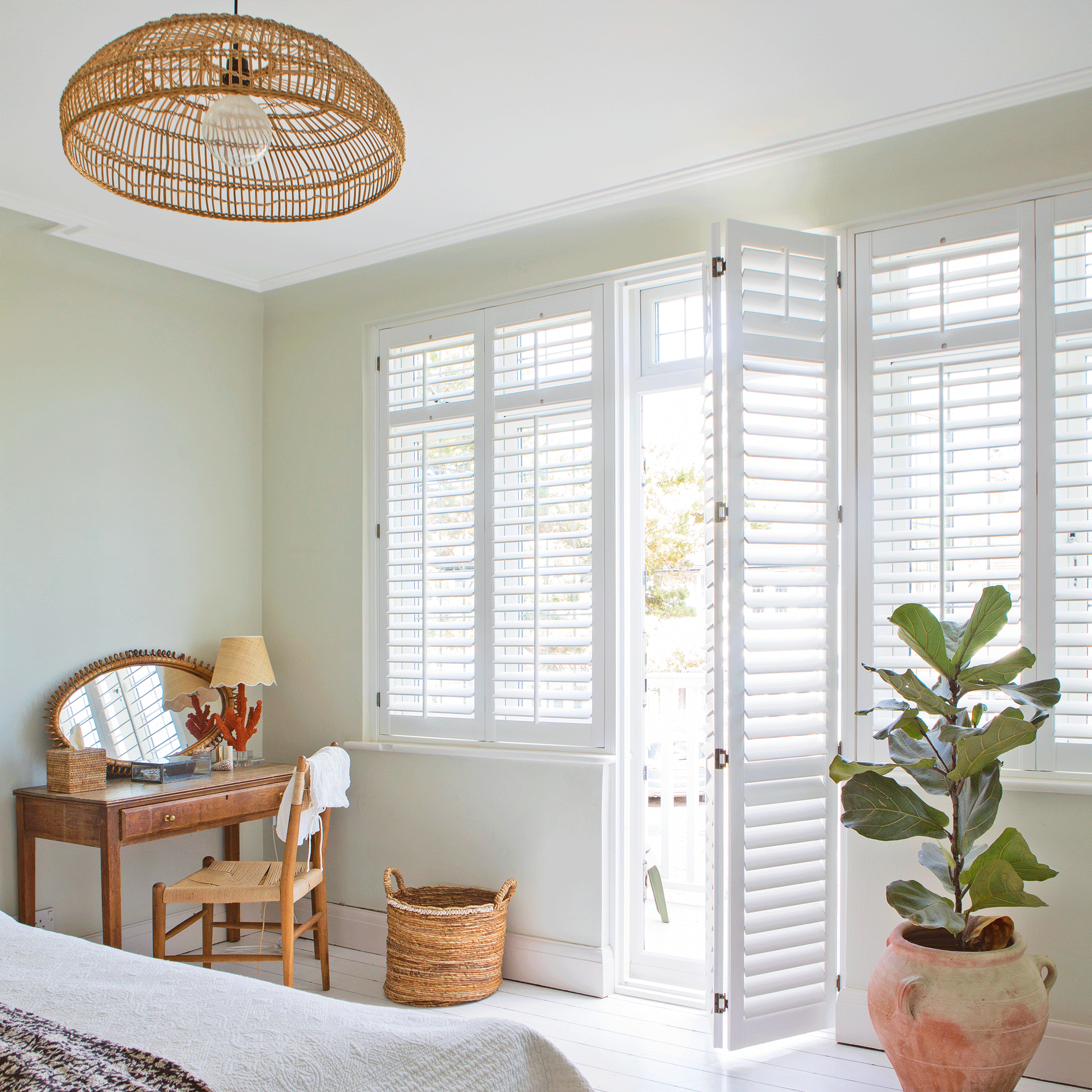a neutral bedroom with white shuttered windows and balcony door, wooden dressing table and a fiddle fig plant