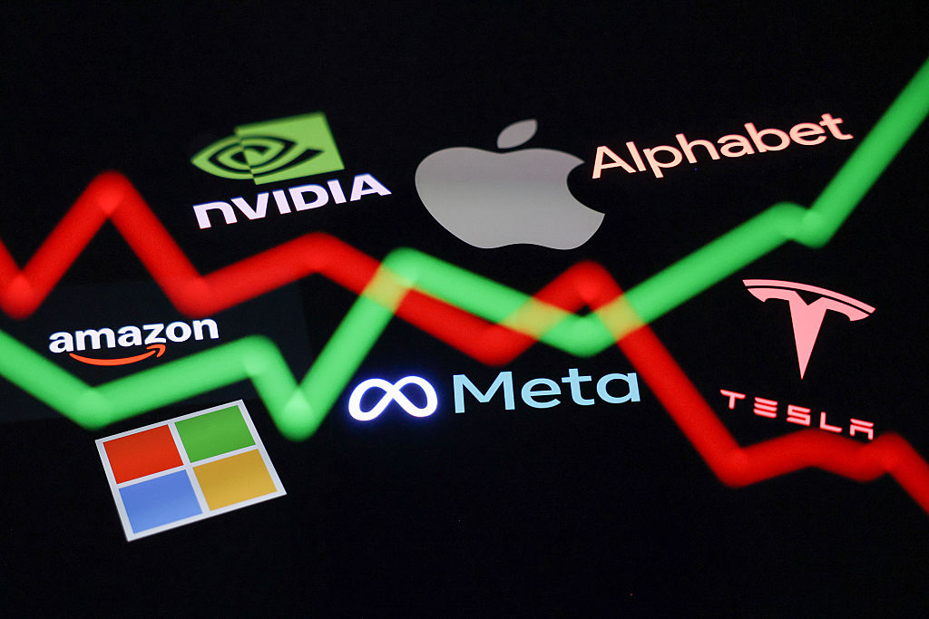 Nvidia, Apple, Alphabet, Amazon, Microsoft, Meta and Tesla logo displayed on a phone screen with stock graph displayed on a laptop screen