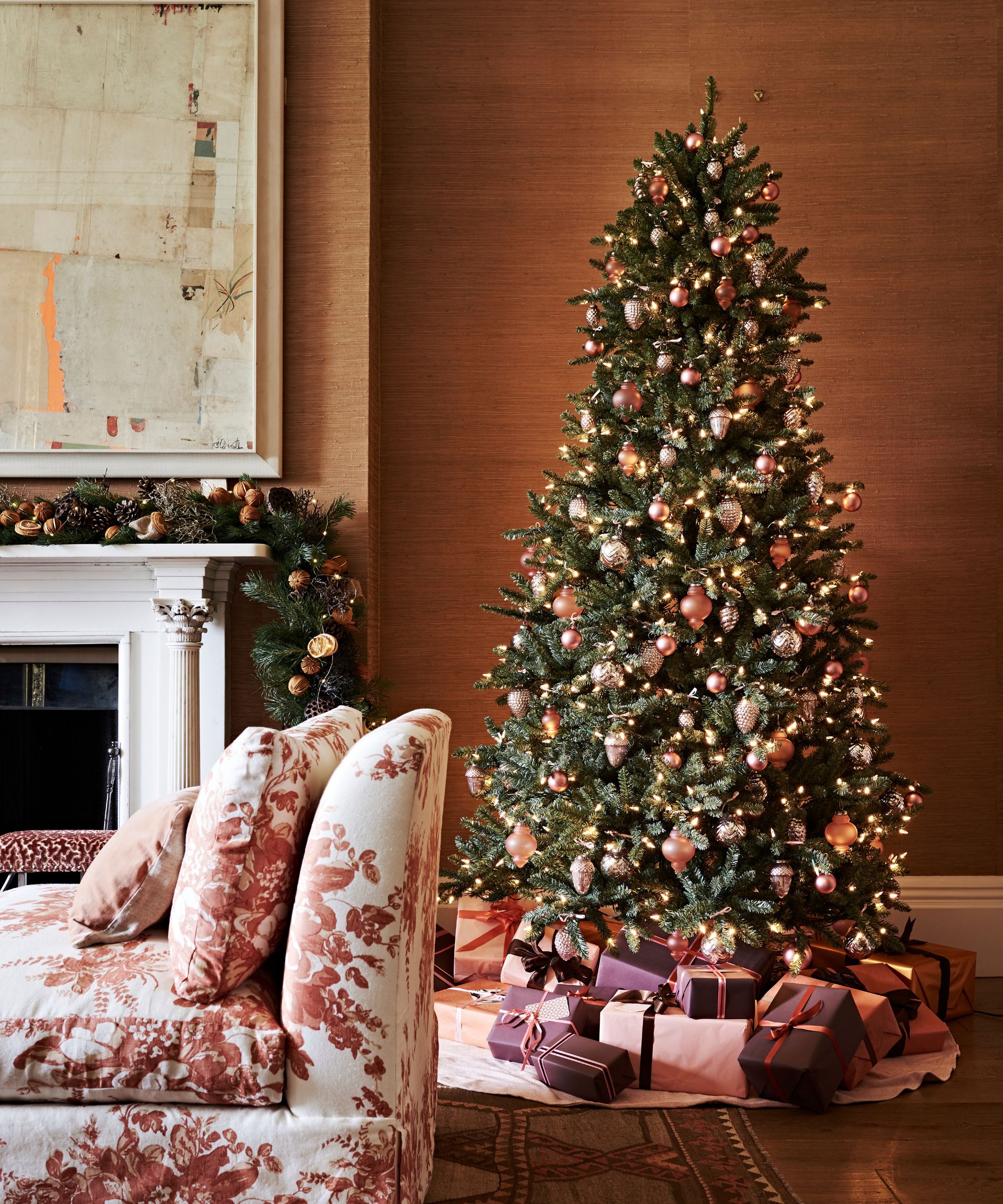 A cozy living room with white and pink floral couch, white mantelpiece with Christmas foliage, large artwork, and large decorated Christmas tree with presents underneath.