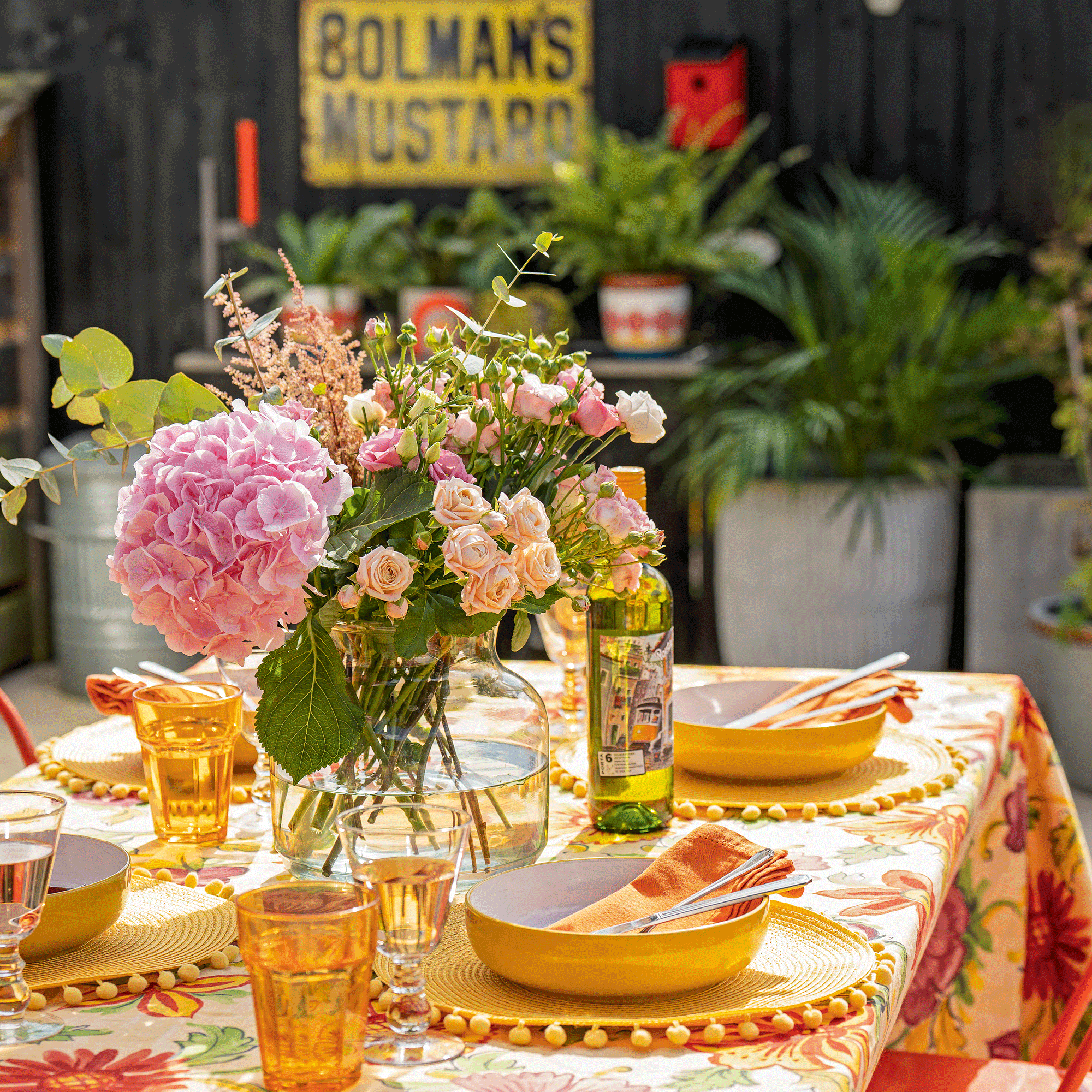 a close up of a dining table in a garden with colourful tablecloth and tableware with a large vase of flowers featuring pink hydrangeas and mini roses