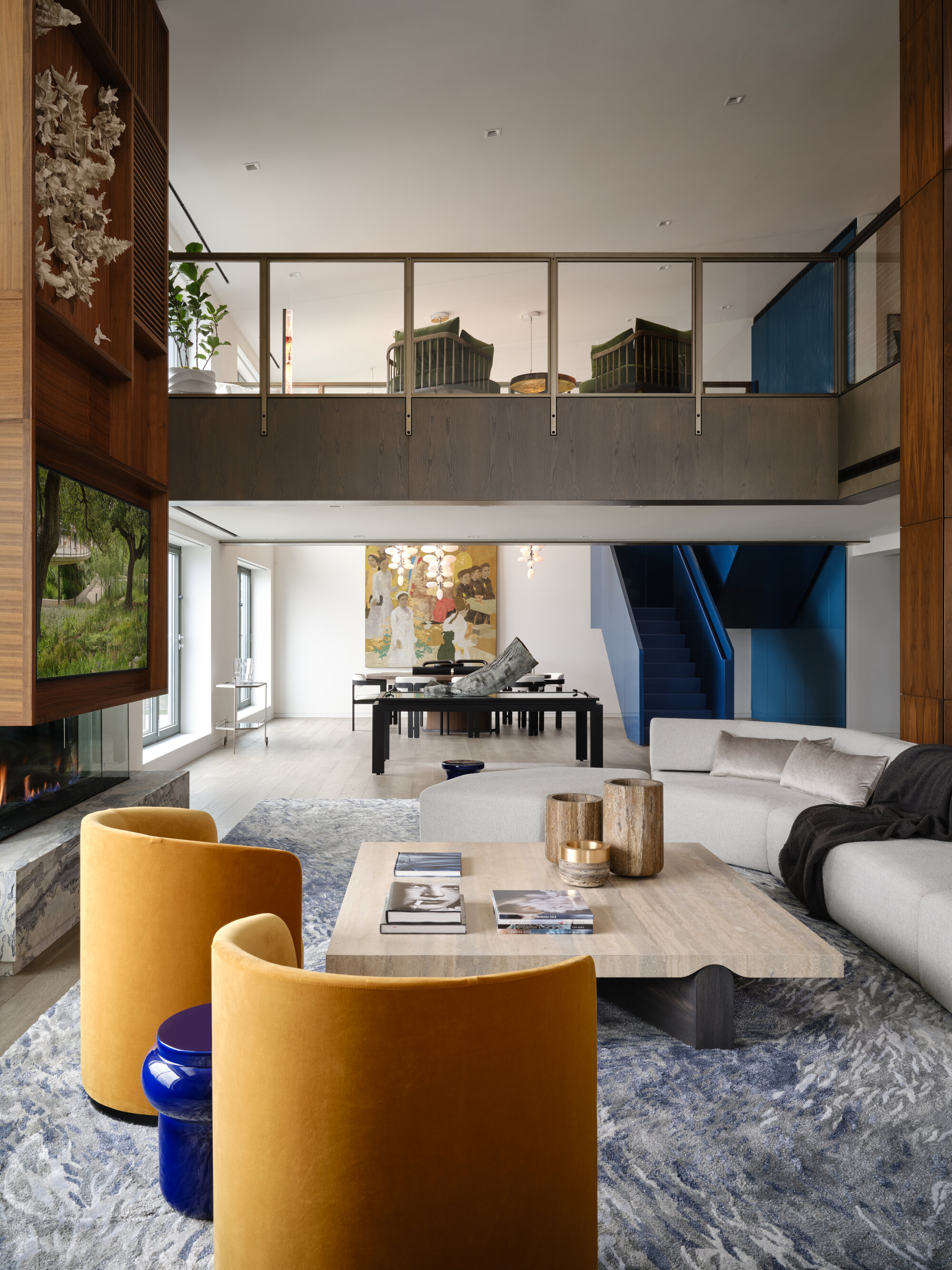 modern living room with double height ceiling, two round yellow armchairs, a shaggy gray rug, large square travertine coffee table and gray sofa. there is a dining area in the distance, and a lattice-clad fireplace with TV