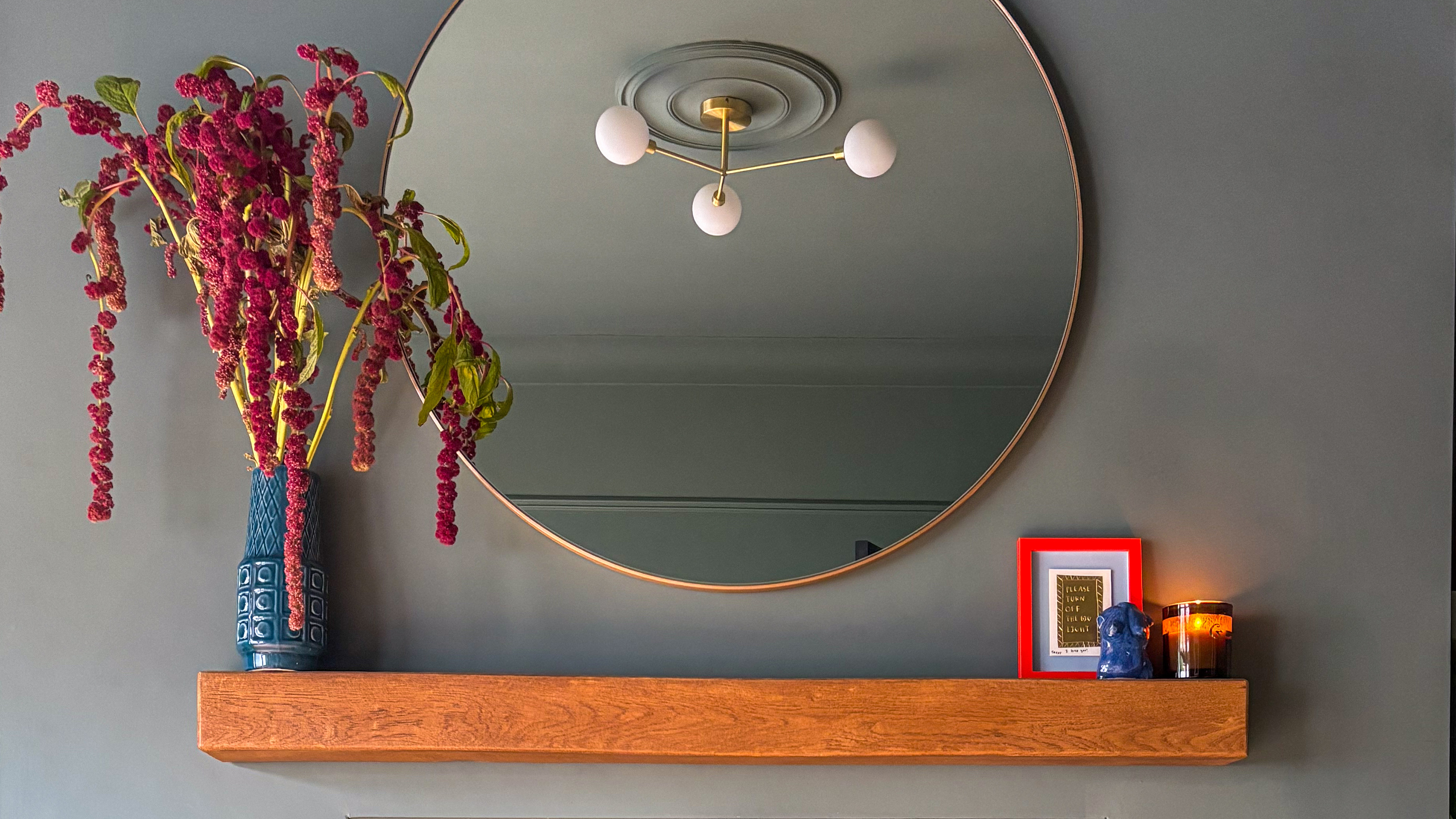 Wooden floating shelf on a grey wall with vintage vase filled with flowers on the left and a modern circular mirror above it