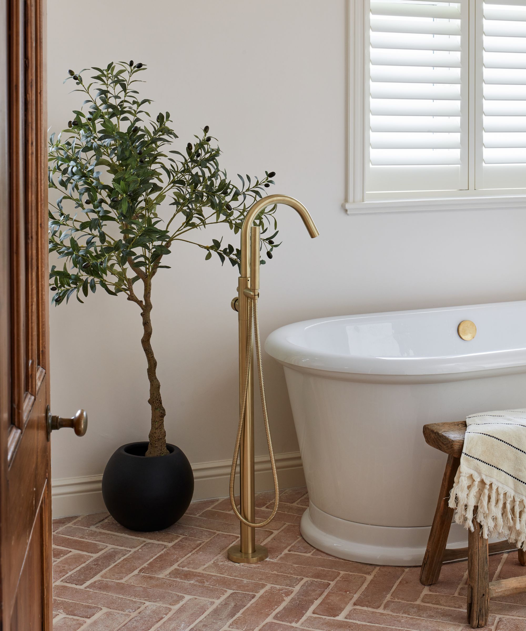 A bathroom with terracotta floor tiles, a white freestanding bath, a brass floor mounted tub filler, and a wooden bath stool