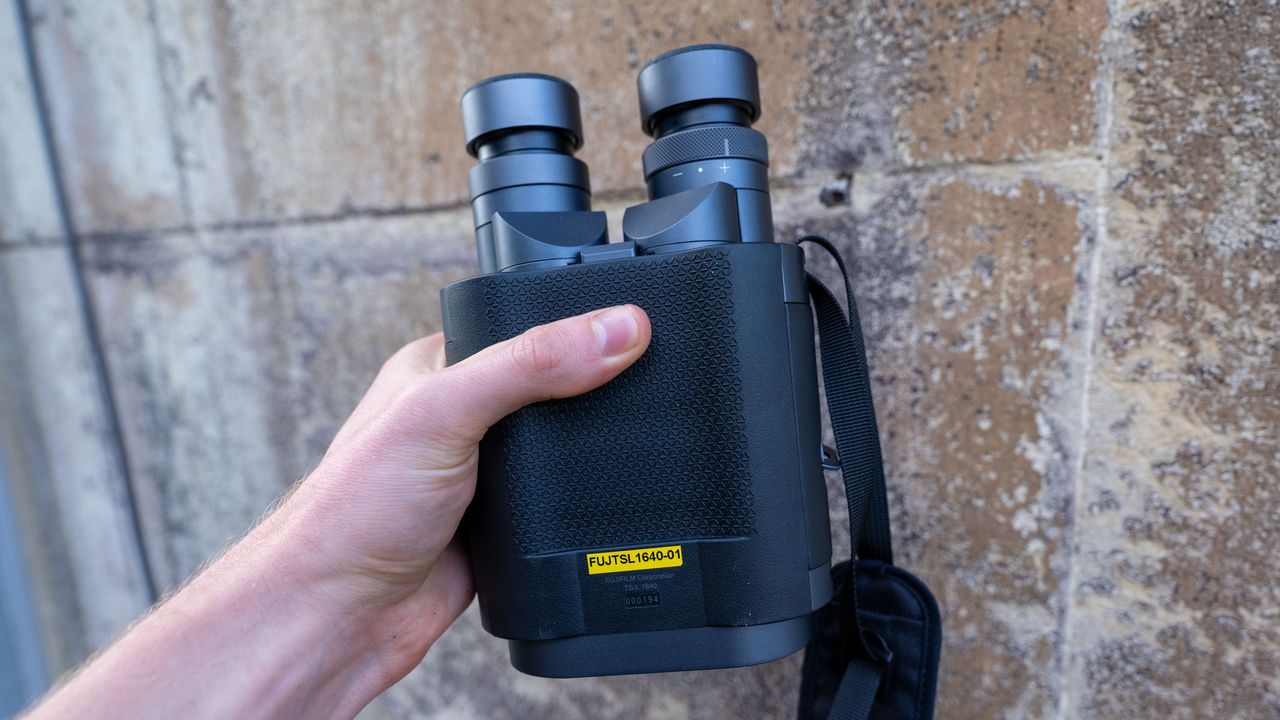 A hand holding the Fujifilm Techno-Stabi TS-L 1640 image-stabilized binoculars with a beige wall in the background.