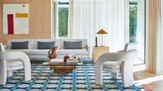 modern living room with white sofa and white armchairs opposite, a timber and glass coffee table, blue patterned rug, timber wall paneling with artwork and white sheer curtains