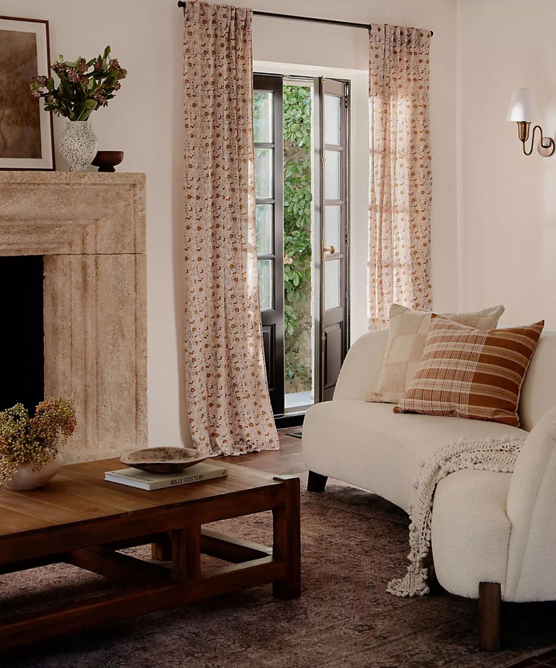 modern rustic timeless living room designed by Amber Lewis for anthropologie with a stone fireplace, dark vintage-style rug, curved modern boucle couch and floral print curtains hung at french doors