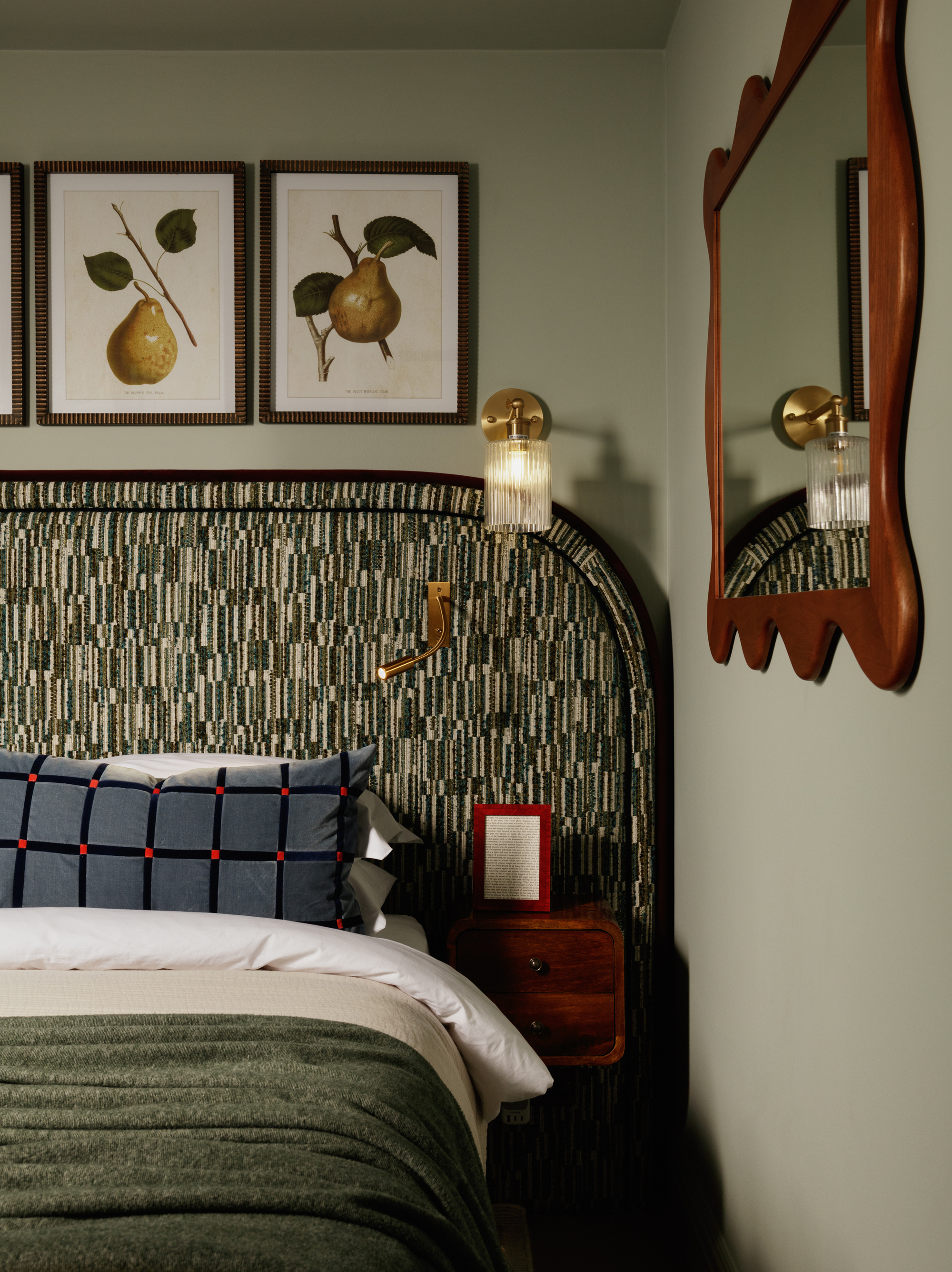 A bedroom with a patterned headboard, a blue checkered bolster pillow, and botanical prints on the wall