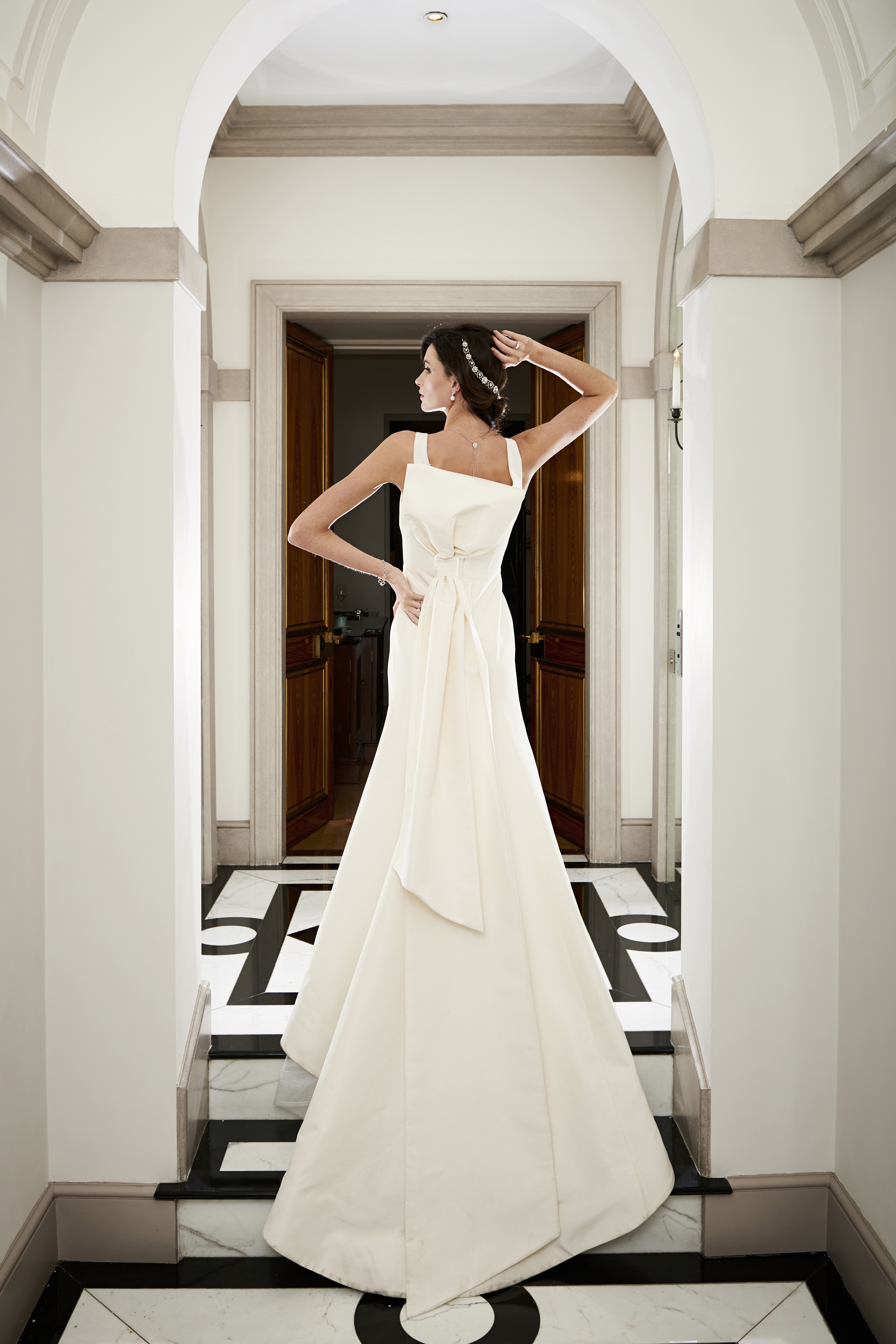 Girl wearing a white wedding dress with a bow, photographed from behind with one arm of her hip and the other on her head