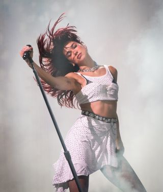 Dua Lipa performing during the Glastonbury Festival on June 28, 24 while wearing a white crop top with a skirt made out of a white skirt. She's holding onto a mic stand and whipping her hair.