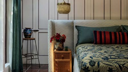 bedroom with floating bed with upholstered bedhead, rattan side tables, tapestry-style bedcover, cabinets behind, stool with vase on top, blue curtain with pendant light hanging down