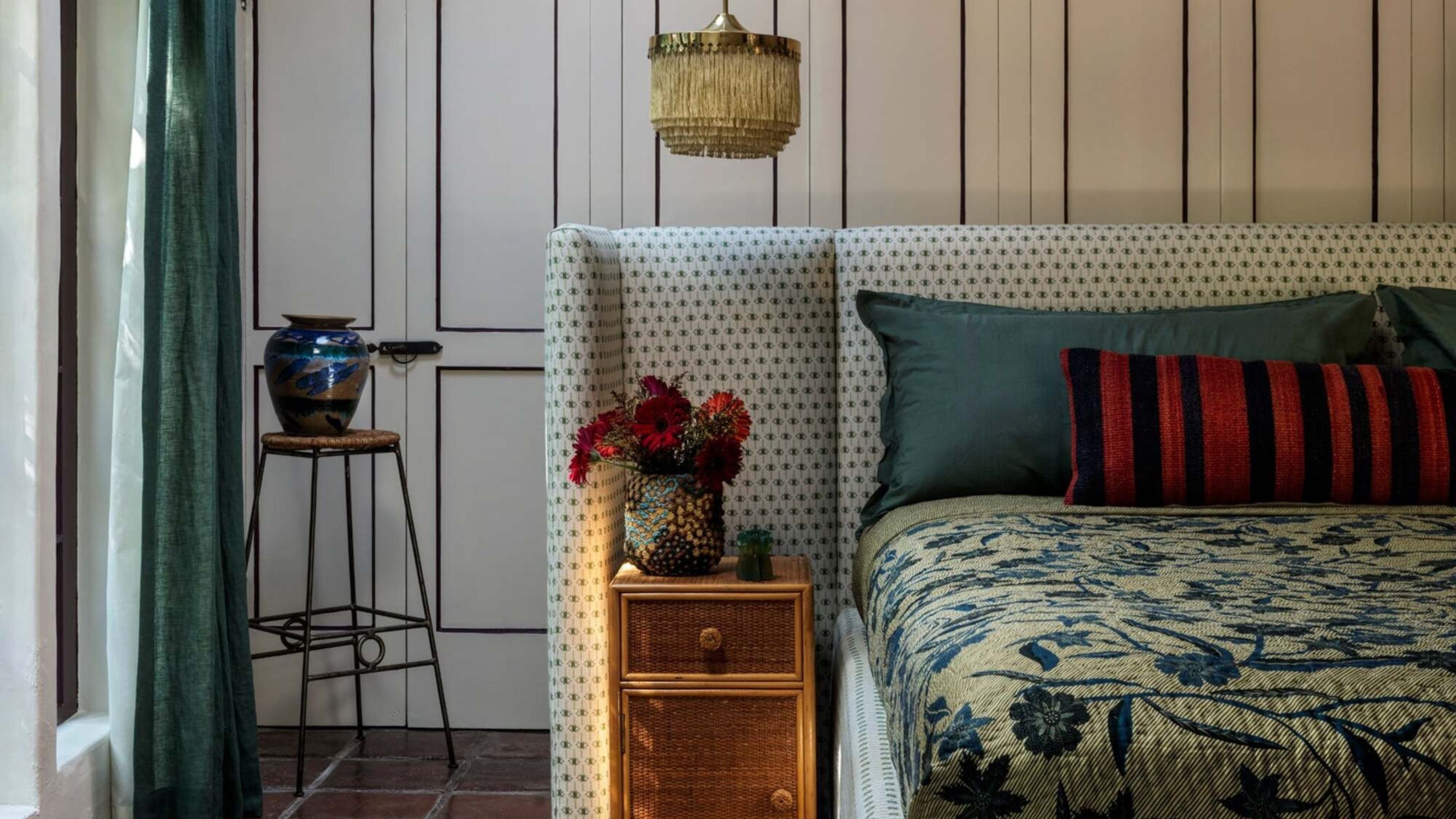 bedroom with floating bed with upholstered bedhead, rattan side tables, tapestry-style bedcover, cabinets behind, stool with vase on top, blue curtain with pendant light hanging down