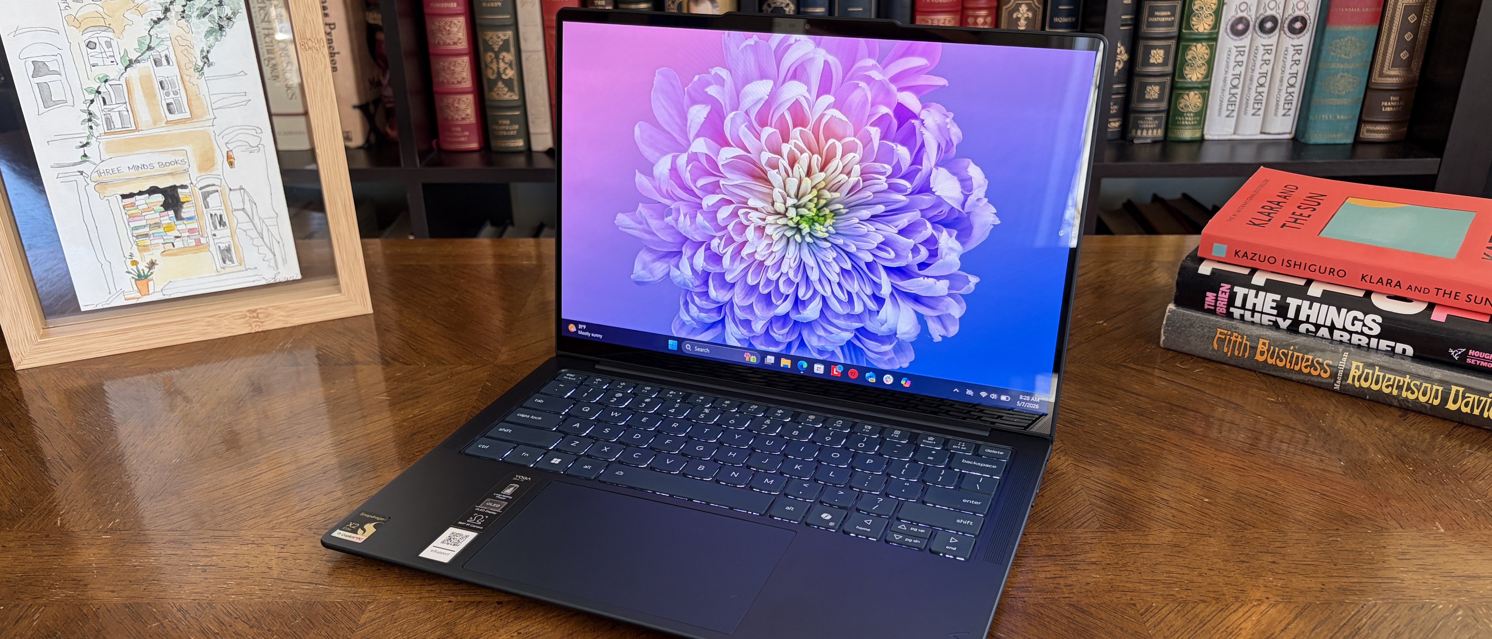 Laptop on wooden desk with a vibrant purple flower wallpaper. Framed art on the left, and a stack of books on the right. Background shows bookshelves.