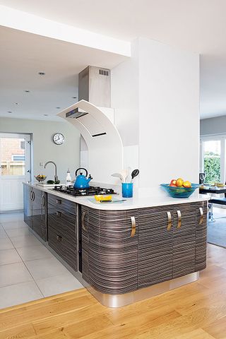 a kitchen extension in a 1960s house, with a kitchen island with grey cabinets, wooden and tiled floor, and a large extractor fan unit