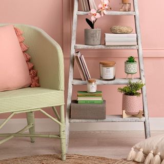 Green chair next to wooden ladder with books, plants and ornaments on wooden floor in front of pink wall