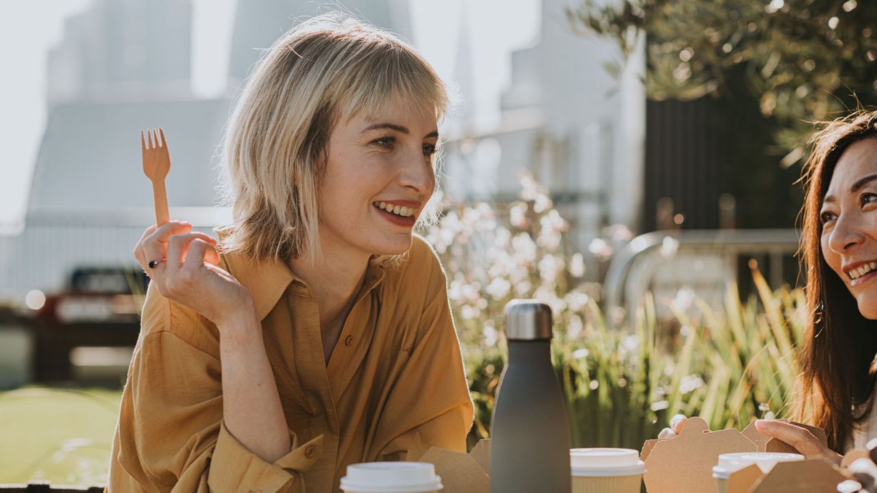 Woman holding up a wooden fork, sitting down at table outdoors with takeaway boxes representing ultra-processed foods, talking with friends