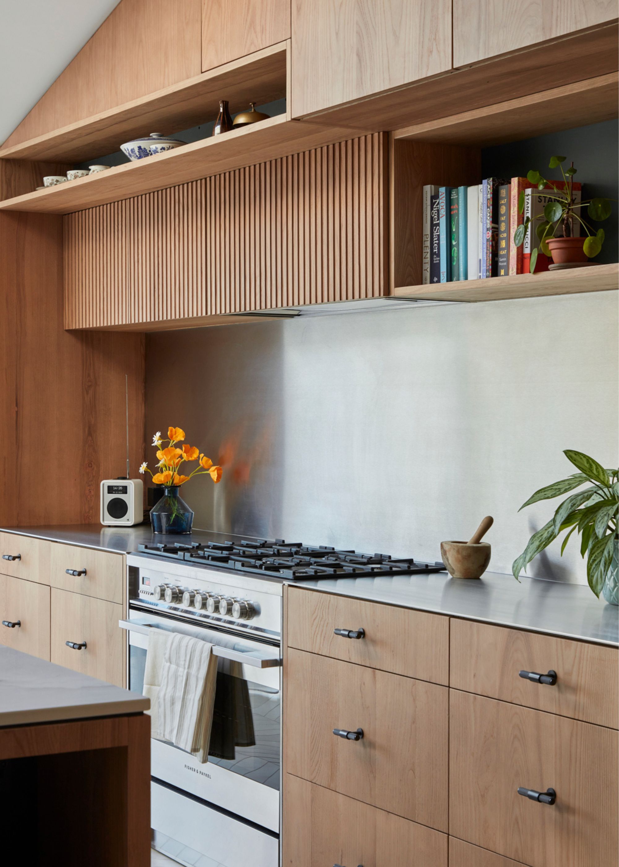 Stainless steel used on a kitchen worktop that also has contrasting oak cabinets and material used around it. There is also a view of the stove beside the stainless steel worktop as well as an oven and shelving above the countertops