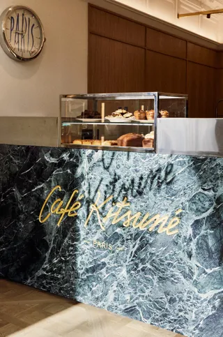 The green marble coffee counter of a bistro-inspired cafe in Paris features an inscribed "Cafe Kitsune" logo in gold. Above it are a pastry display in glass and a vintage-looking clock that reads "Paris"