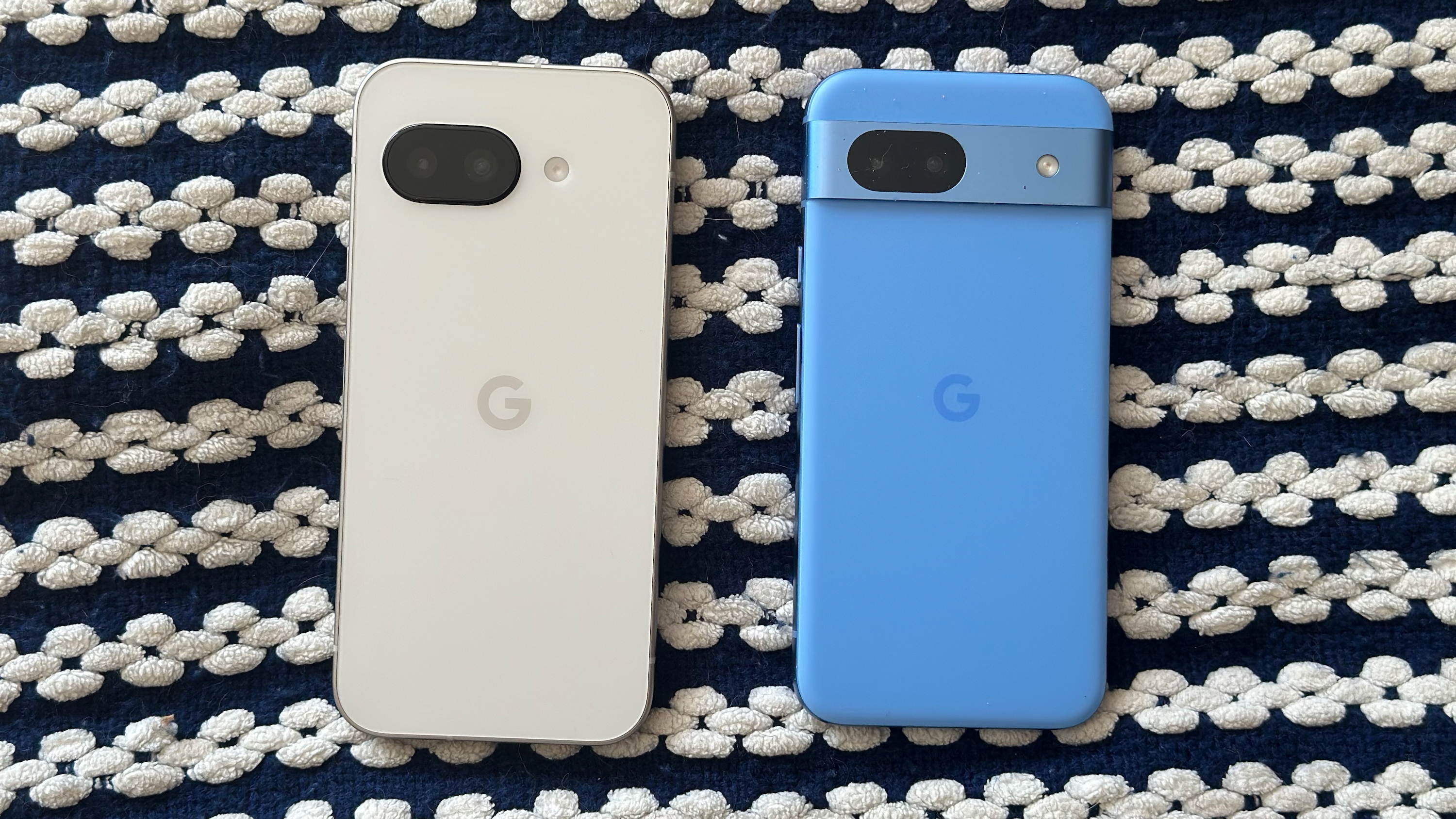 The Google Pixel 9a (left) and Google Pixel 8a (right) laying on a blanket face-down, showing the 9a's new flat camera cutout and larger, flatter design compared to the more rounded 8a with a metal camera bar.