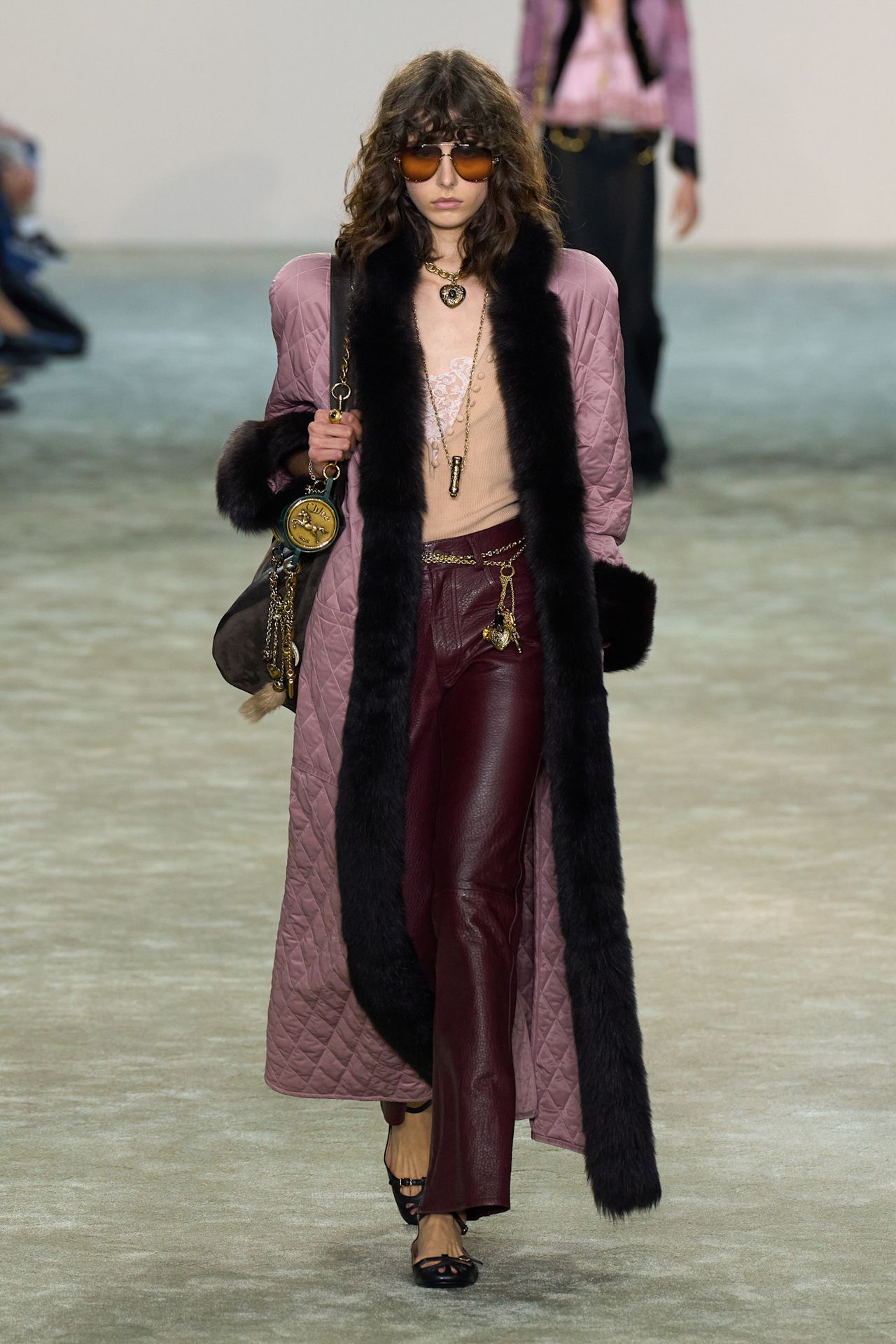 A model walking in the Chlo&eacute; F/W 25 runway show wearing a mauve quilted coat with fur trim, leather pants, and a tan low-cut top.