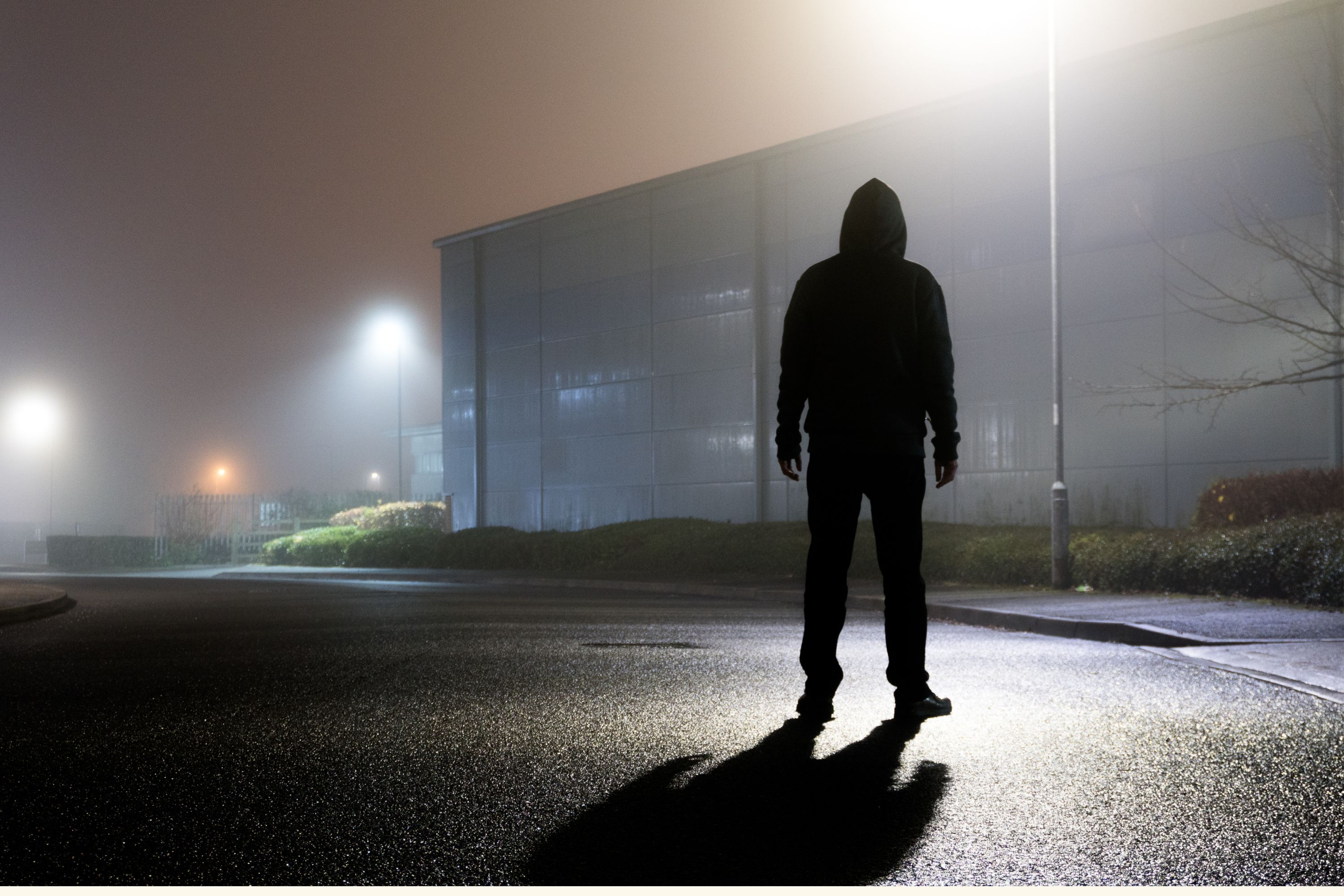 A sinister hooded figure, silhouetted by street lights on an empty industrial estate on a mysterious foggy night