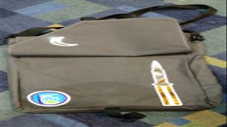 The International Astronautical Congress 2019 conference bag on display in the Aerothreads booth, set against a rendering of an employee painting of the James Webb Space Telescope.