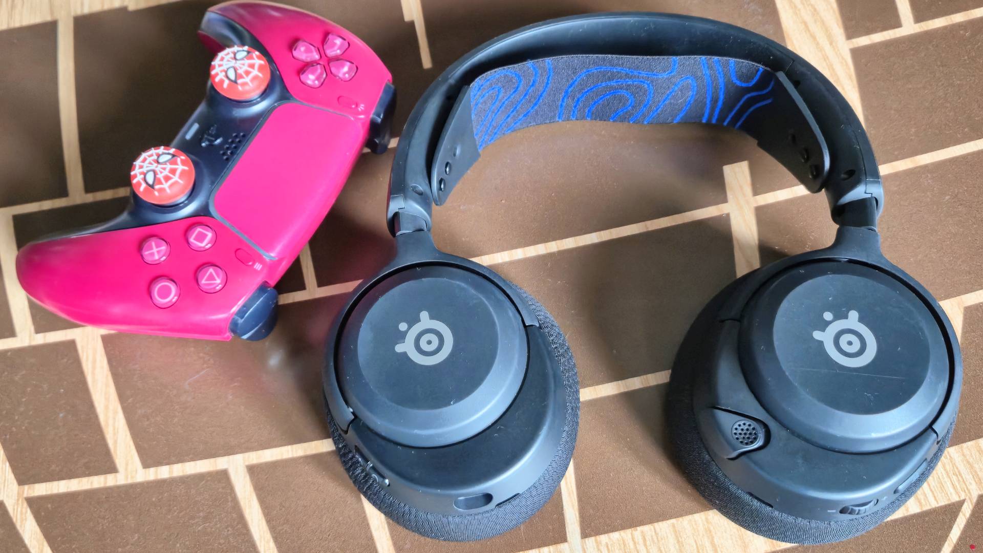 Photo by Rosalie Newcombe of the SteelSeries Arctis Nova 5P headset sitting on a wooden table with a red DualSense controller sitting to its left.